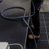 A person wearing tan work boots and dark trousers stands on Fine Ribbed Rubber Matting in an industrial setting. A blue hose is coiled nearby on the ribbed black surface.