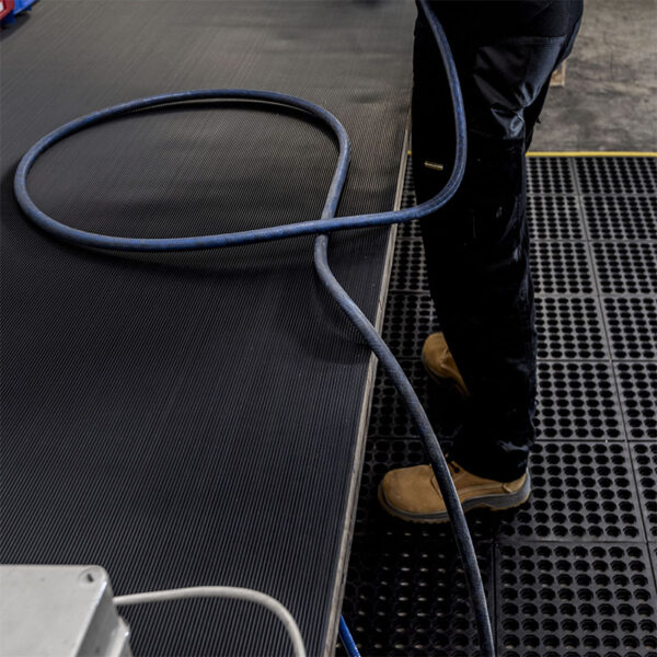 A person wearing tan work boots and dark trousers stands on Fine Ribbed Rubber Matting in an industrial setting. A blue hose is coiled nearby on the ribbed black surface.