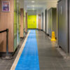 A corridor in a changing room features light green and wooden lockers, a swimming pool mat running down the centre, and a yellow caution wet floor sign near the showers.