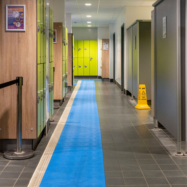 A corridor in a changing room features light green and wooden lockers, a swimming pool mat running down the centre, and a yellow caution wet floor sign near the showers.