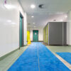 A clean, well-lit changing room features green and grey cubicles, teal doors, tiled floors, and a blue swimming pool mat. A yellow caution sign stands at the back.