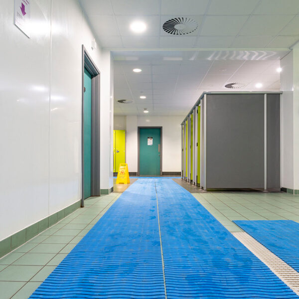 A clean, well-lit changing room features green and grey cubicles, teal doors, tiled floors, and a blue swimming pool mat. A yellow caution sign stands at the back.