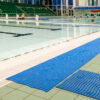 An indoor swimming pool features blue Swimming Pool Mats along the tiled poolside, clear water in empty lanes, and green spectator seats in the background.