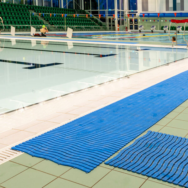 An indoor swimming pool features blue Swimming Pool Mats along the tiled poolside, clear water in empty lanes, and green spectator seats in the background.