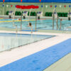 The Swimming Pool Mat is shown in an indoor pool with blue lane dividers, metal ladders, and a lifeguard chair. Swimmers use the pool as green seats and triangular flags are visible in the background.