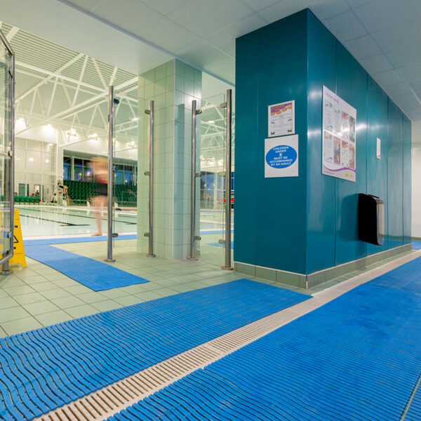 An indoor swimming pool area features blue Swimming Pool Mats on the floor, glass doors, a green tiled column, and informational posters on a teal wall. The pool and seating area can be seen in the background.
