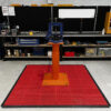 A metal tool or press on an orange stand sits centred on a Multi-Purpose Interlocking PVC Tile in a workshop, with shelves of tools, equipment, and storage bins visible in the background.
