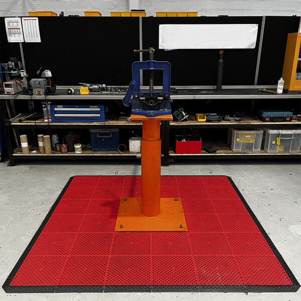 A metal tool or press on an orange stand sits centred on a Multi-Purpose Interlocking PVC Tile in a workshop, with shelves of tools, equipment, and storage bins visible in the background.