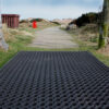 The Playground Safety Mat is a large black rubber mat with circular holes, shown outdoors on a wooden path, surrounded by grass, bushes, and a distant red box under cloudy skies.