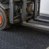 Close-up of a Forklift Mat with a forklift’s front wheel and metal forks resting on its textured black surface, situated inside an industrial warehouse with a white wall in the background.