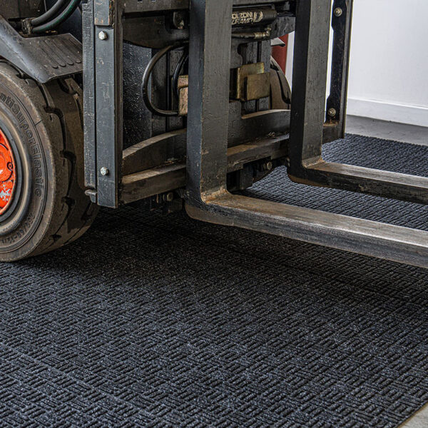 Close-up of a Forklift Mat with a forklift’s front wheel and metal forks resting on its textured black surface, situated inside an industrial warehouse with a white wall in the background.