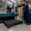 A worker stands on an Interlocking Anti-Fatigue Mat while operating heavy machinery in a brightly lit factory, surrounded by metal structures and equipment.