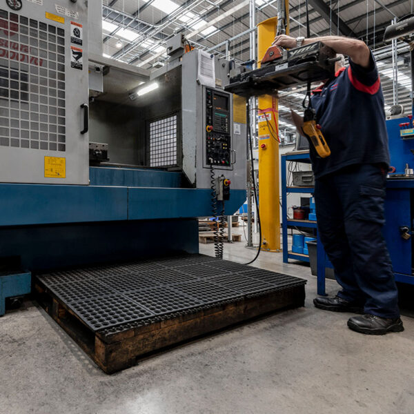 A worker stands on an Interlocking Anti-Fatigue Mat while operating heavy machinery in a brightly lit factory, surrounded by metal structures and equipment.