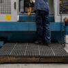 A person in blue work clothes operates a large blue and grey industrial machine, standing on an interlocking anti-fatigue mat in a factory.