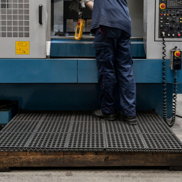 A person in blue work clothes operates a large blue and grey industrial machine, standing on an interlocking anti-fatigue mat in a factory.