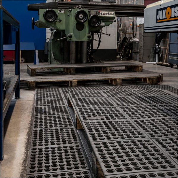A factory floor features Interlocking Anti-Fatigue Mats alongside a large green industrial drilling machine, with pallets stacked below and various other workplace equipment visible.