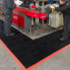 Two workers operate industrial machinery in a workshop, standing on Custom Oil Resistant Mats with interlocking black tiles and red borders, placed on the concrete floor around the equipment.