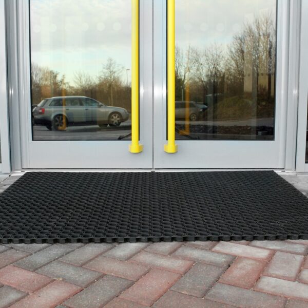 An Octagonal Ring Mat in black rubber is placed outside a glass door with two bright yellow handles. A car is parked nearby, and trees can be seen behind the red and grey brick paving.