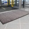 A High-Performance Entrance Mat in grey is placed on a tiled floor at the entrance of a building with glass doors, with cars and a car park visible outside.
