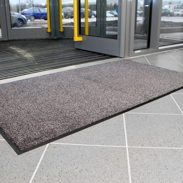 A High-Performance Entrance Mat in grey is placed on a tiled floor at the entrance of a building with glass doors, with cars and a car park visible outside.