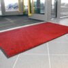 A High-Performance Entrance Mat in bright red is positioned just inside glass doors on light grey tiles, with cars visible outside.