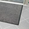 The High-Performance Entrance Mat, featuring a grey textured surface with a black border, is shown on a light grey tiled floor near a glass door.