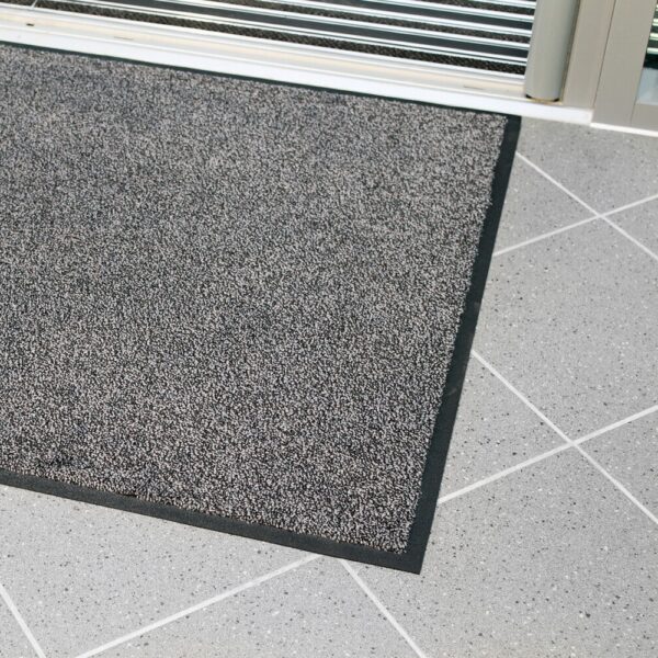 The High-Performance Entrance Mat, featuring a grey textured surface with a black border, is shown on a light grey tiled floor near a glass door.