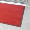 The High-Performance Entrance Mat, featuring a red surface with a black border, is positioned on a light grey tiled floor by a doorway with a metallic threshold.
