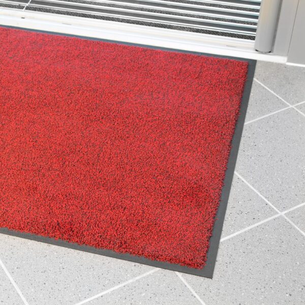 The High-Performance Entrance Mat, featuring a red surface with a black border, is positioned on a light grey tiled floor by a doorway with a metallic threshold.