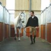 A man in riding gear leads a white horse by a headcollar inside a stable with wooden and metal walls, featuring the Amoebic Top Stable Mat.