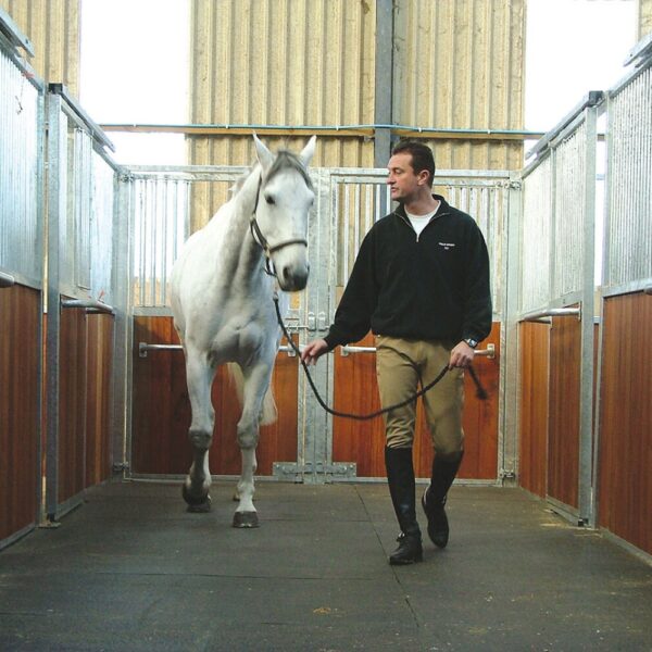 A man in riding gear leads a white horse by a headcollar inside a stable with wooden and metal walls, featuring the Amoebic Top Stable Mat.