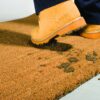 A person in yellow steel toe boots steps onto the Weather-Resistant Entrance Mat, leaving muddy footprints on its brown surface.