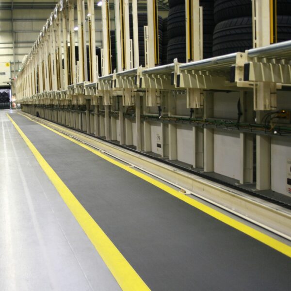 Rows of stacked tyres in an industrial warehouse, featuring a Standard Anti-Fatigue Mat with a yellow-striped walkway running alongside automated equipment and storage shelves.