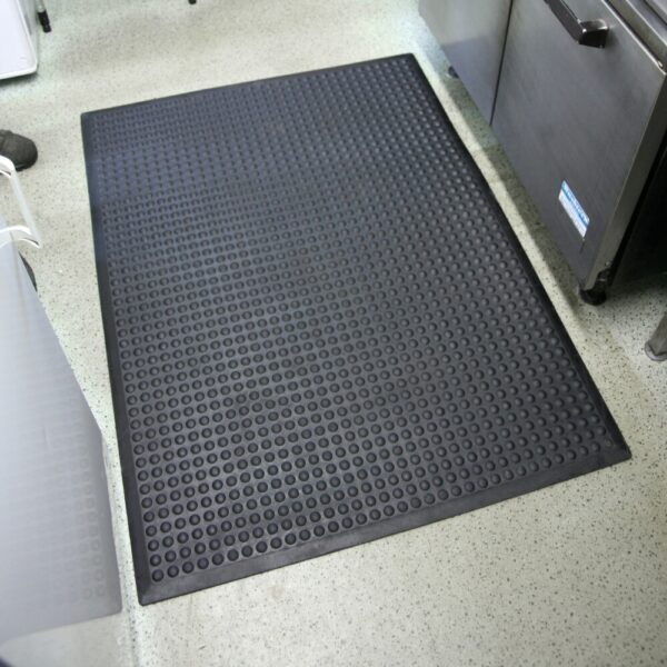 An Air Cushioned Workshop Mat with a black raised circular pattern is placed on a light speckled floor in a commercial kitchen, surrounded by stainless steel equipment.