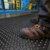 Close-up of a person wearing worn brown work shoes with laces, standing on the Air Cushioned Workshop Mat with raised bumps in an industrial setting with blue and yellow equipment in the background.