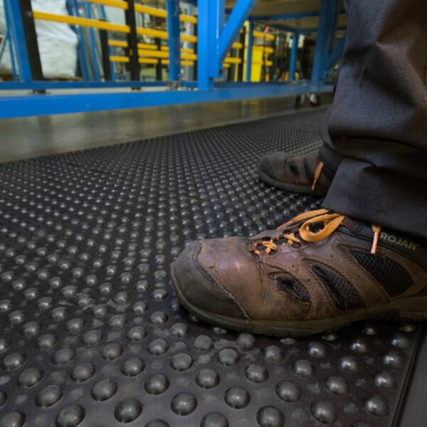 Close-up of a person wearing worn brown work shoes with laces, standing on the Air Cushioned Workshop Mat with raised bumps in an industrial setting with blue and yellow equipment in the background.