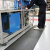 A person in dark clothing operates a large blue and silver machine in a clean, well-lit factory, standing on an Interlocking Air Cushioned Mat with a textured black surface.
