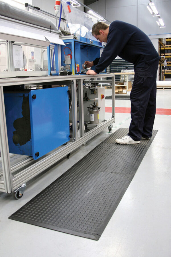 A person in dark clothing operates a large blue and silver machine in a clean, well-lit factory, standing on an Interlocking Air Cushioned Mat with a textured black surface.