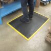 A person wearing black shoes and blue trousers stands on an Air Cushioned Workshop Mat with a yellow border in a workshop, next to blue shelving and a trolley.