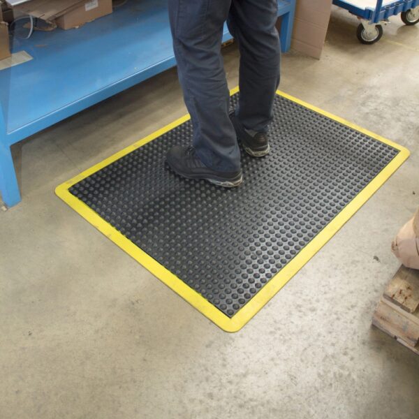 A person wearing black shoes and blue trousers stands on an Air Cushioned Workshop Mat with a yellow border in a workshop, next to blue shelving and a trolley.