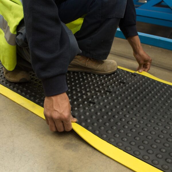 A person in a high-visibility vest and work shoes is kneeling on concrete, installing or adjusting an Interlocking Air Cushioned Mat with black surface and yellow edges.