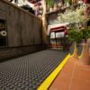 A close-up of a flower shop floor features the Air Cushioned Workshop Mat with a yellow edge on tiled flooring, surrounded by metal flower buckets, a wooden counter, and stacked chairs against the wall.