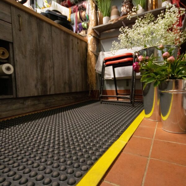 A close-up of a flower shop floor features the Air Cushioned Workshop Mat with a yellow edge on tiled flooring, surrounded by metal flower buckets, a wooden counter, and stacked chairs against the wall.