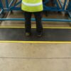 A person wearing dark trousers and a high-visibility yellow vest stands on the Air Cushioned Workshop Mat, which is black with yellow edges, in an industrial setting. Only their lower half is visible.