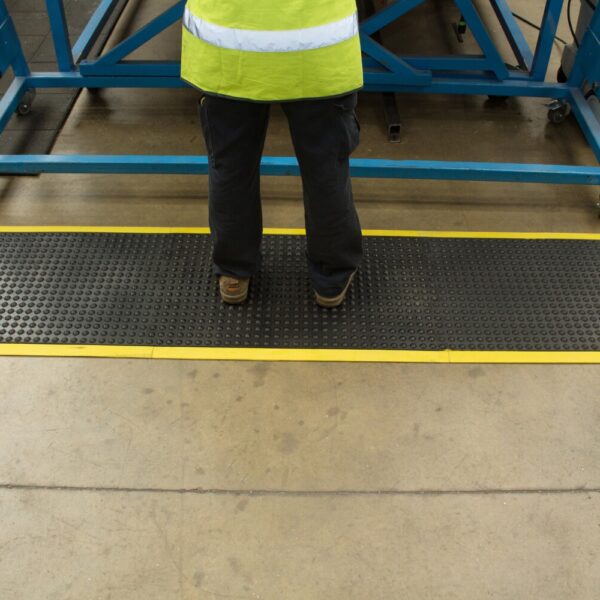 A person wearing dark trousers and a high-visibility yellow vest stands on the Air Cushioned Workshop Mat, which is black with yellow edges, in an industrial setting. Only their lower half is visible.