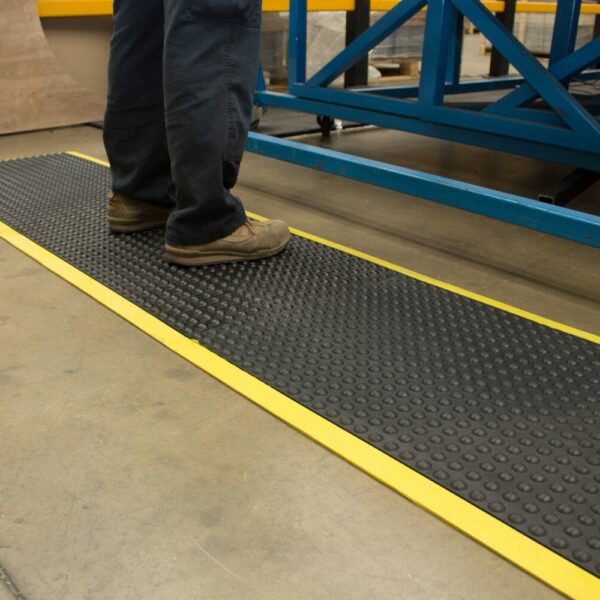 A person in work trousers and tan shoes stands on the Interlocking Air Cushioned Mat, which is black with yellow edges, in an industrial setting.