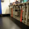 A workshop features metal workbenches, orange coiled air hoses, tools on surfaces, and the floor covered with Circular Stud Rubber Matting for anti-fatigue comfort in an organised, industrial setting.