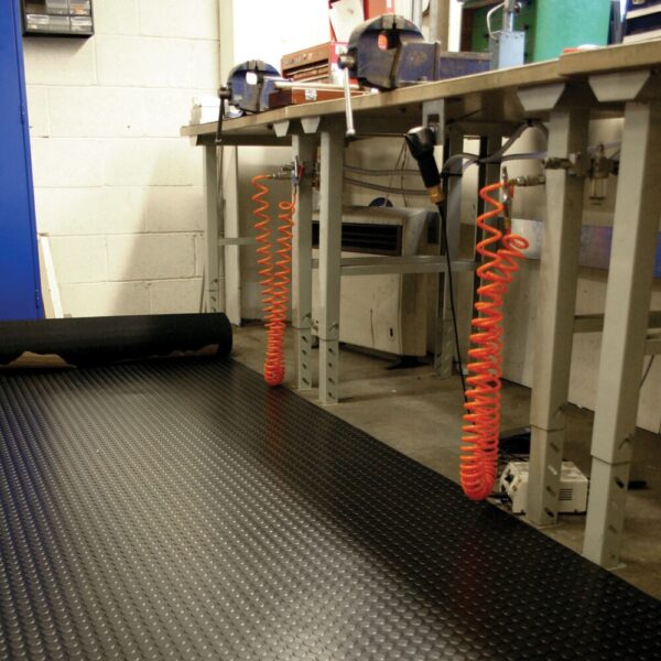 A workshop features metal workbenches, orange coiled air hoses, tools on surfaces, and the floor covered with Circular Stud Rubber Matting for anti-fatigue comfort in an organised, industrial setting.
