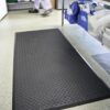The Deckplate Anti-Fatigue Mat in black is placed on a speckled floor in a commercial kitchen, with shelves of crockery on the right and a person walking in the background.