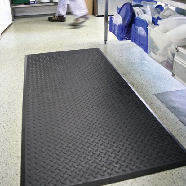 The Deckplate Anti-Fatigue Mat in black is placed on a speckled floor in a commercial kitchen, with shelves of crockery on the right and a person walking in the background.